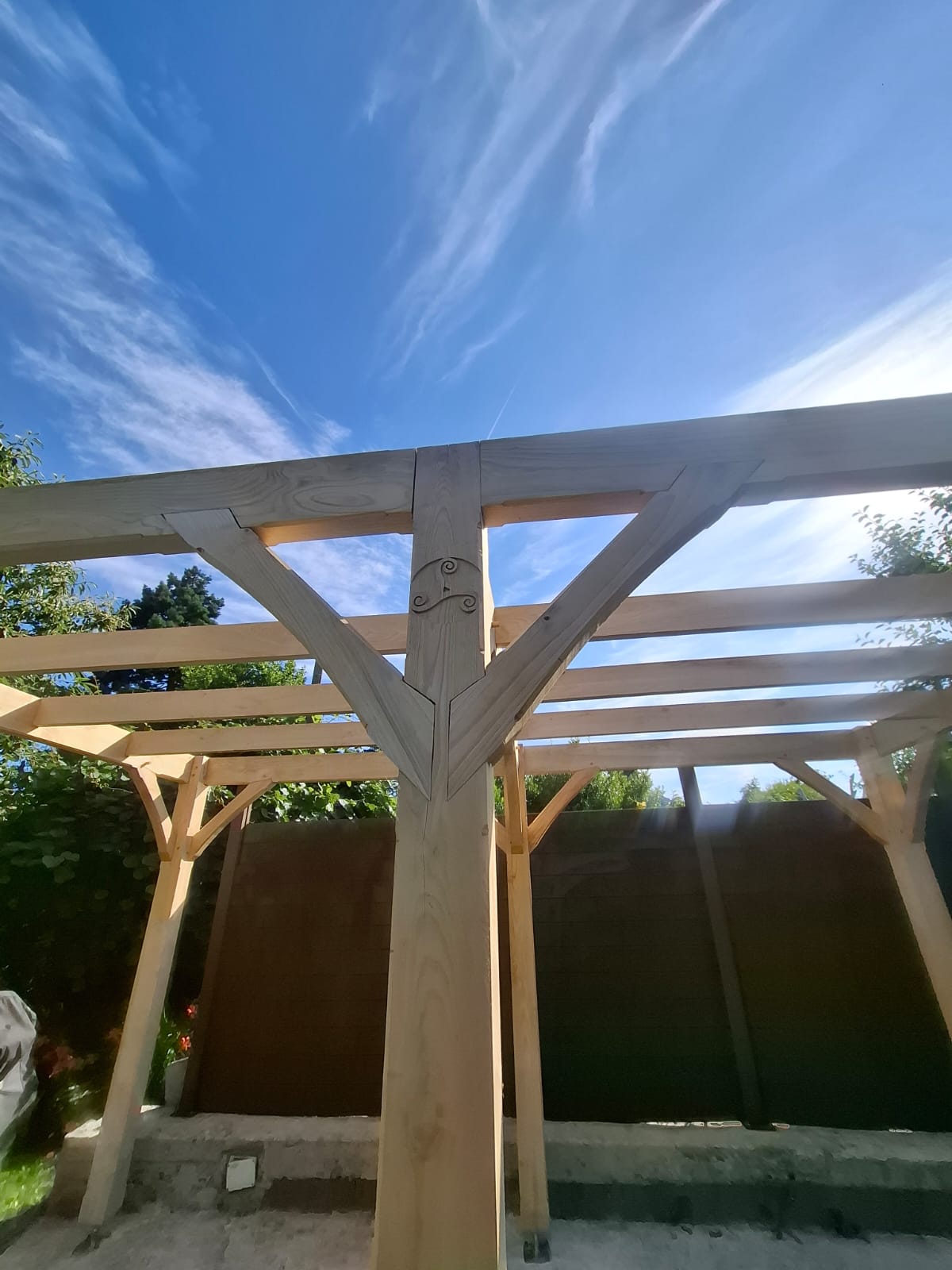 Pergola en châtaignier en contre-plongée, assemblage central et triskell sous le ciel bleu